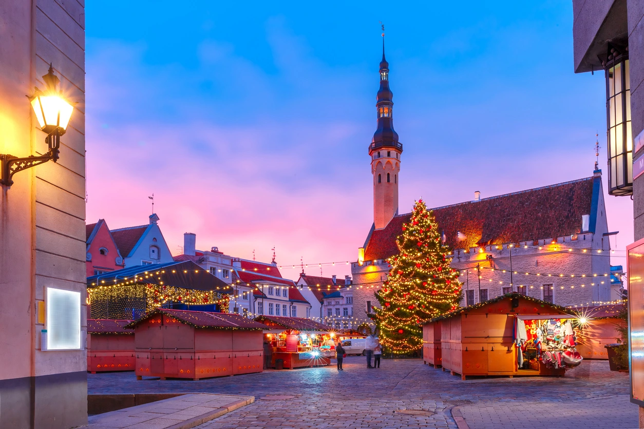 Tallinn Eski Şehir Meydanı'nda, mavi ve pembe tonlarındaki alacakaranlık gökyüzünün altında, ışıklarla süslenmiş büyük bir Noel ağacı, tarihi belediye binasının sivri kulesi ve etrafında kurulu ahşap Noel pazarı tezgahları, sıcak bir şenlik atmosferi yaratıyor.
