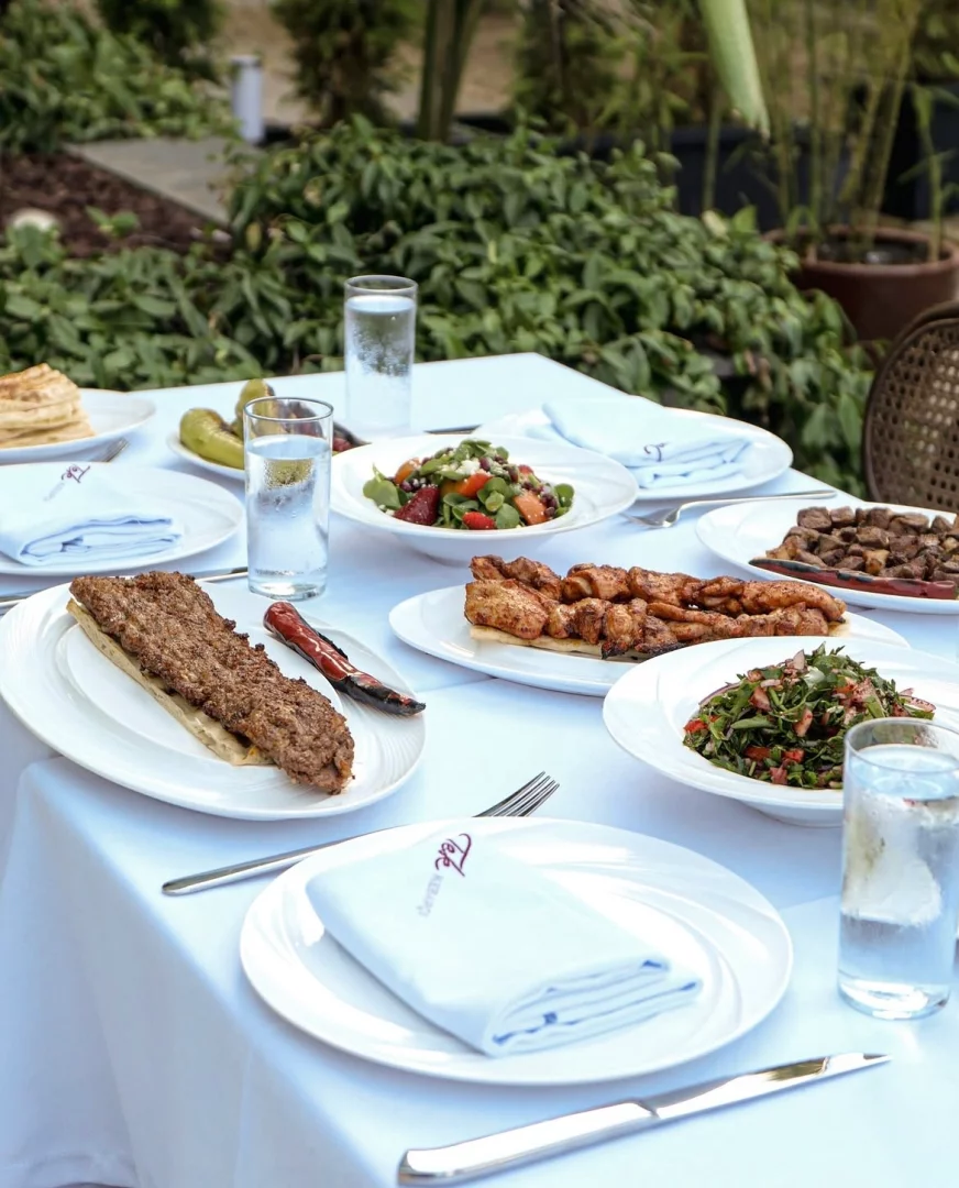 Tek Kebapçı Etiler'deki açık hava restoranında, beyaz örtülü bir masada uzun Adana kebap, tavuk şiş, doğranmış et, zengin salatalar ve turşularla dolu geleneksel bir Türk ziyafeti sunumu, yemyeşil bahçe ortamında iştah açıcı bir şekilde sergileniyor.