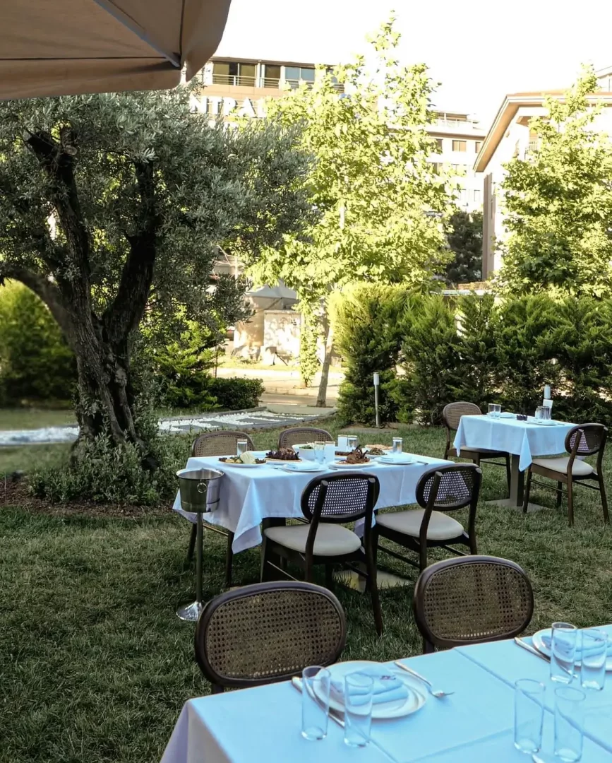 Etiler'deki Tekkebapçı restoranının zeytin ağaçları ve yeşil bitki örtüsüyle çevrili bahçesinde, beyaz masa örtüleri serilmiş ve üzerinde tabaklar ile bardaklar bulunan, bazı masalarda yiyeceklerin de yer aldığı, misafirlerini bekleyen şık ve ferah bir açık hava yemek alanı.