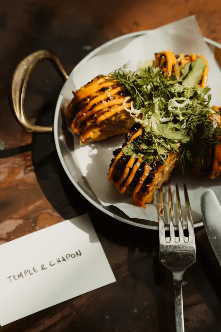 Temple & Chapon’da, New York chophouse ve Fransız mutfağı sentezini yansıtan menüden; üzerine parlak turuncu sos gezdirilmiş, taze yeşilliklerle zenginleştirilmiş dilimlenmiş ızgara bir yemeğin, metal bir servis tavasında sunulduğu an, yanında çatal-bıçak ve restoranın kartı.