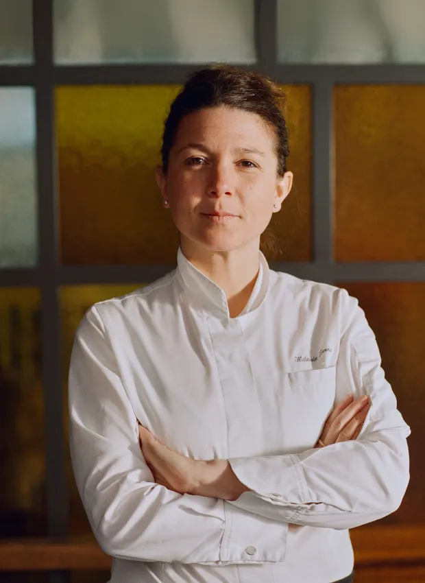 Temple & Chapon'un danışman şefi Mélanie Serre, beyaz şef ceketiyle kolları kavuşmuş, kendine güvenli bir ifadeyle kameraya bakıyor; arkasındaki sıcak tonlardaki cam detaylar, New York chophouse ve Fransız mutfağını birleştiren modern restoranın etkileyici atmosferini vurguluyor.