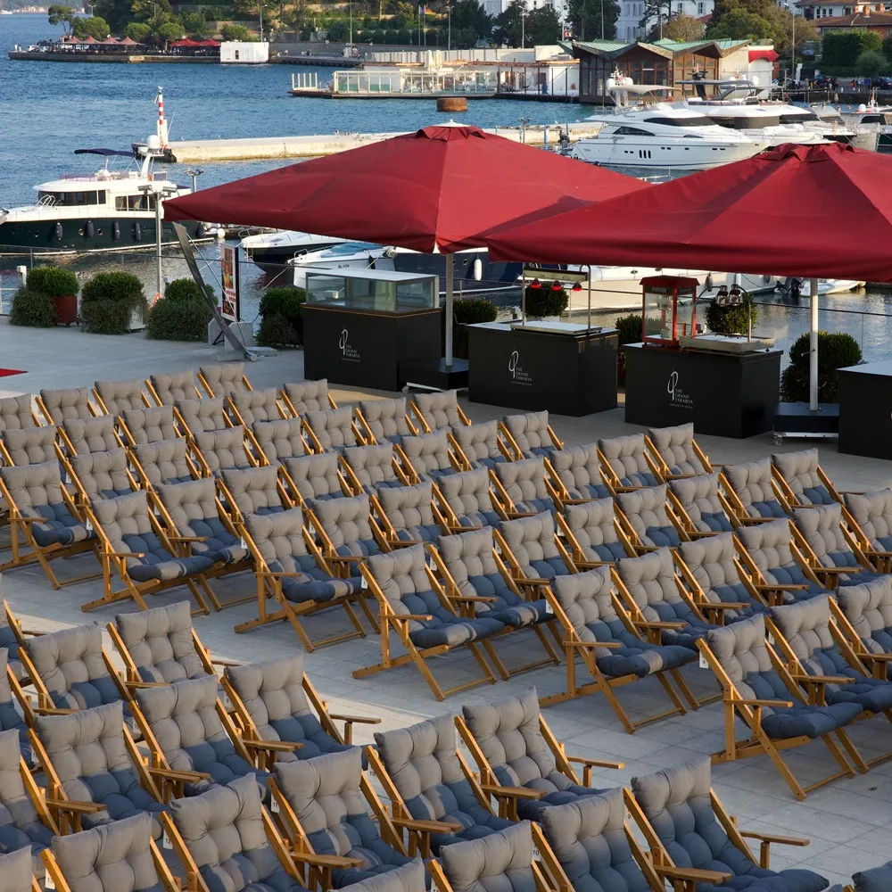 The Grand Tarabya'nın Bosphorus'a nazır terasında, açık hava sineması etkinliği için sıralanmış yüzlerce gri minderli ahşap şezlong, arkasında kırmızı şemsiyeler altında ikram standları ve lüks yatların demirli olduğu deniz manzarası.