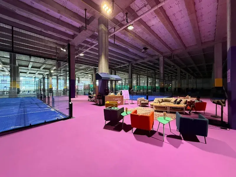 İstanbul'daki The Padel House tesisinde, mor tonlarında aydınlatılmış beton tavanlı geniş bir alanda, parlak fuşya zeminin solunda mavi padel kortları ve ortada modern koltuk takımlarının yer aldığı dinlenme alanı görülüyor.