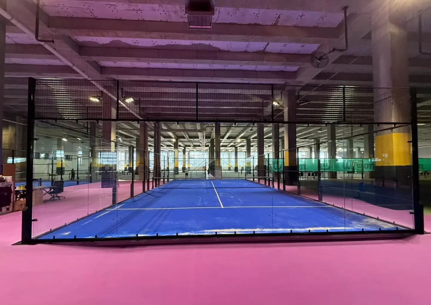Padel House İstanbul tesisindeki geniş ve modern bir salonda, pembe zeminle çevrili, mavi kaplamalı padel kortu cam ve metal çitlerle sınırlanmış, arkada mor ışıklarla aydınlatılmış diğer kortlar da uzanıyor.