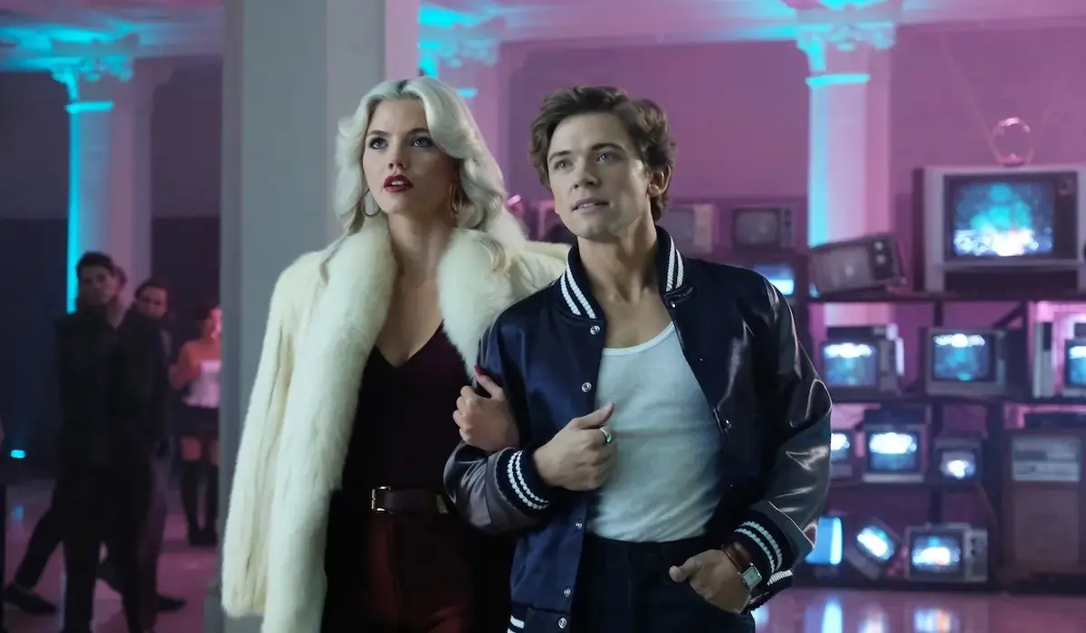 The Shards dizisi sahnesinde, sarışın, hacimli saçlı ve beyaz kürklü bir kadın ile mavi kolej ceketi giyen bir adam, neon mor ve mavi ışıklarla aydınlatılmış, vintage televizyonlarla dolu bir odada dikkatle yukarıya bakıyor.
