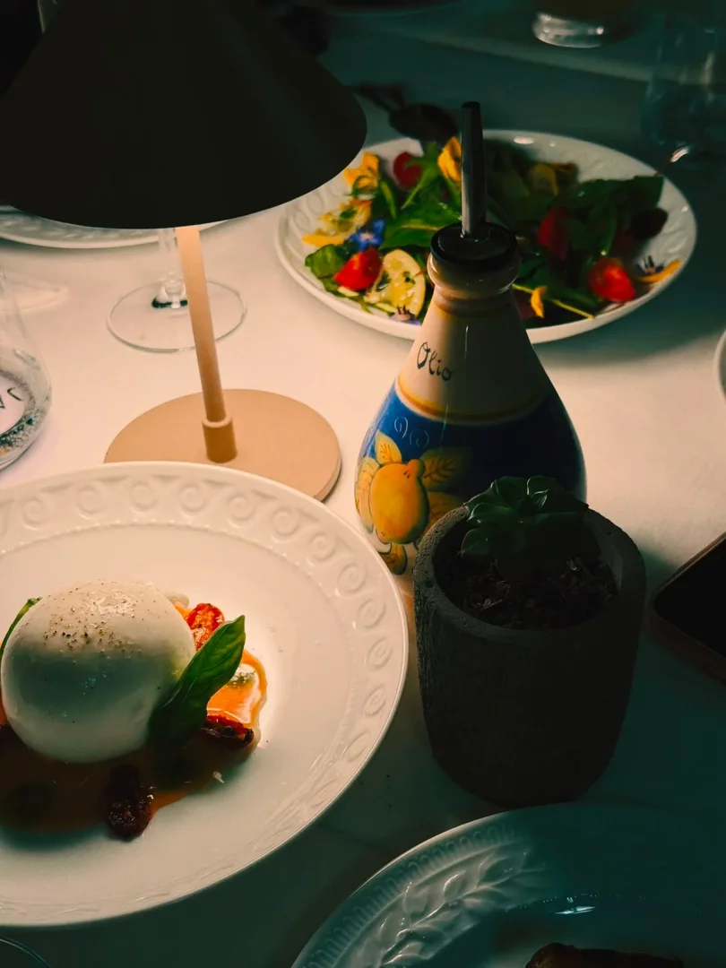 Yarı karanlık bir trattoria masasında, baharatlı burrata peyniri, fesleğen ve kurutulmuş domatesli turuncu sosla servis edilmiş bir tabağın yanı sıra, arkada parlak bir masa lambası, Olio yazılı zeytinyağı şişesi ve çeşitli renkli sebzelerden oluşan bir salata tabağı, İtalyan yemeklerinin samimi atmosferini vurguluyor.