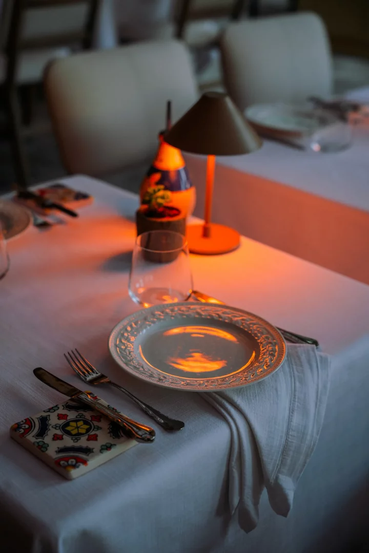 İtalyan trattoria'sında akşam yemeği için beyaz masa örtüsüyle özenle düzenlenmiş, oymalı beyaz bir tabağın, parlak çatal bıçak takımının, boş bir şarap kadehinin, dekoratif seramik altlığın ve sıcak turuncu ışık yayan bir masa lambasının oluşturduğu davetkar masa düzeni.