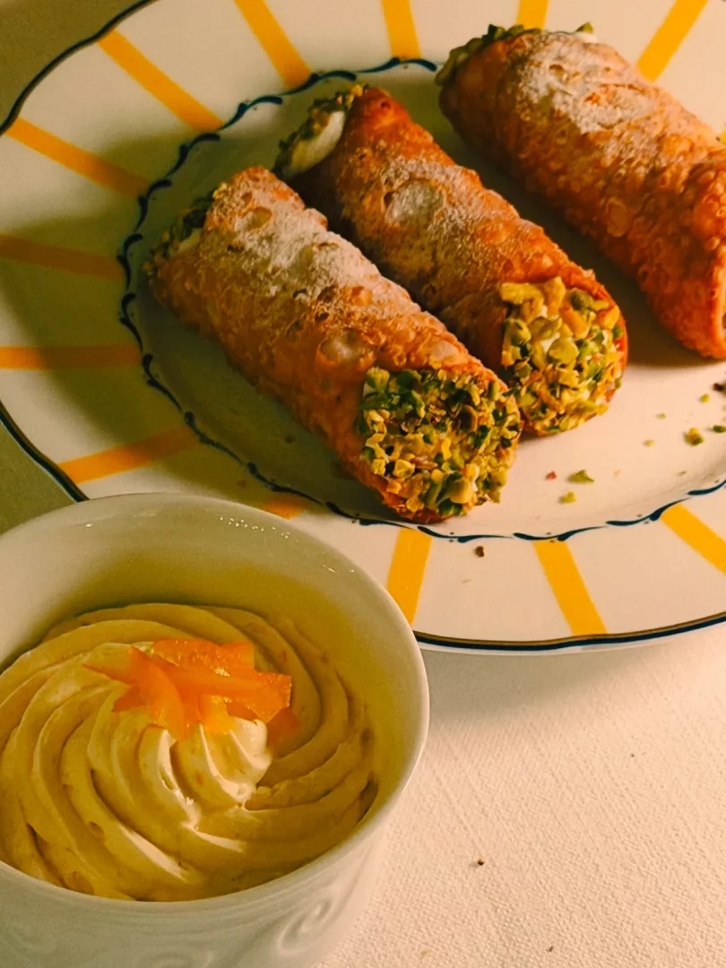 Sarı radyal desenli ve mavi kenarlı beyaz bir tabakta, pudra şekeri serpilmiş ve uçları bol antep fıstığıyla kaplı üç adet çıtır İtalyan cannoli görülüyor; ön planda ise, sarmal desende sıkılmış, üzeri taze portakal zestleriyle süslenmiş kremalı bir tatlı, bu lezzetli trattoria menüsünü tamamlıyor.