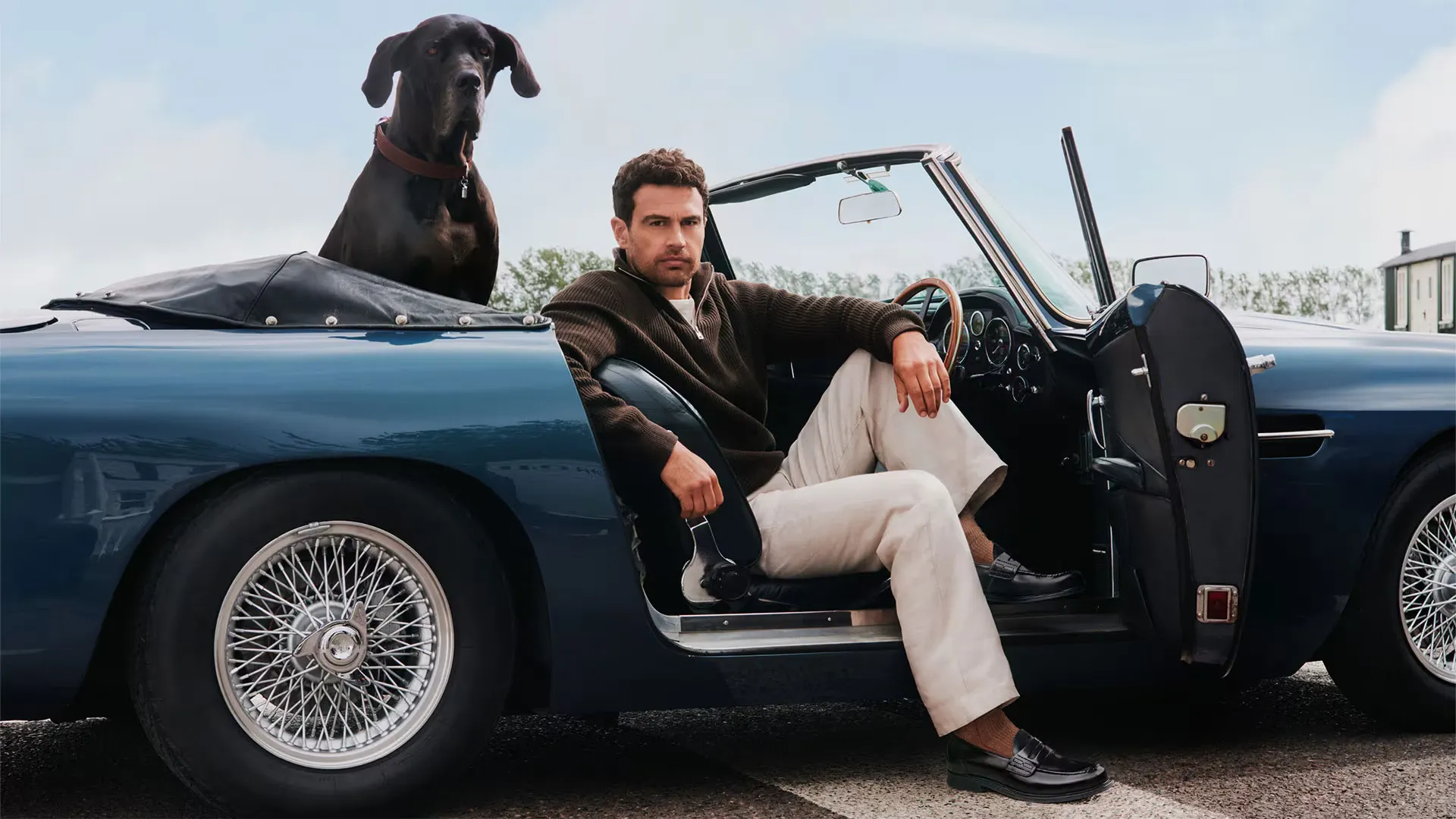 Koyu mavi klasik üstü açık arabasının sürücü koltuğunda oturan Theo James, kahverengi kazak ve açık renk pantolonuyla, bir ayağı dışarıda siyah deri loafer ayakkabısını gösterirken, arka koltukta büyük siyah bir köpekle objektife bakıyor.
