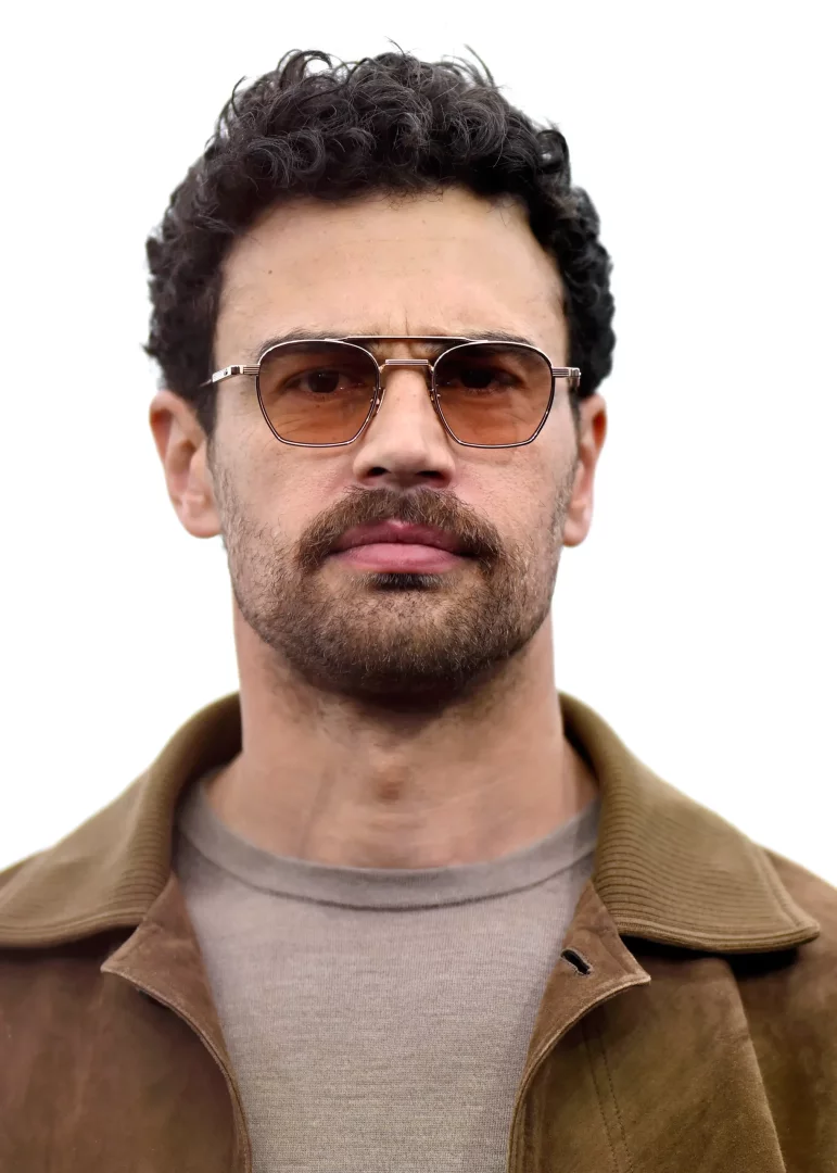 Theo James, kıvırcık koyu saçları, sakal ve bıyığı, kahverengi tonlarında camlara sahip modern metal çerçeveli gözlükleriyle doğrudan kameraya bakarken, kahverengi süet ceketi ve açık renk içliğiyle bu portrede oldukça şık görünüyor.