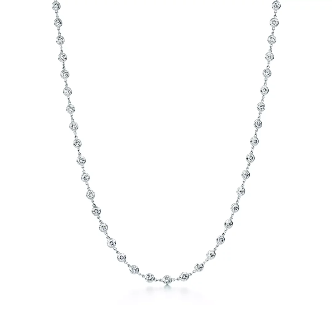 Beyaz bir arka plan üzerinde, zarif ve ince bir zincire eşit aralıklarla yerleştirilmiş çok sayıda yuvarlak pırlanta taşı ile bezeli, ikonik Tiffany Diamonds by the Yard kolyesinin göz alıcı ışıltısını sergileyen bir görüntü.