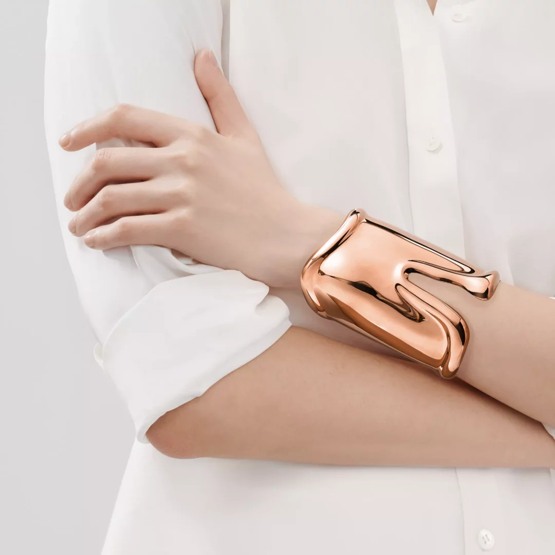 Tiffany Elsa Peretti Bone Cuff bileklik, beyaz uzun kollu gömlek giymiş bir modelin çapraz duran sol kolunda, parlak rose gold rengiyle parlayarak kendine özgü, akışkan ve kemik benzeri formunu sergiliyor.