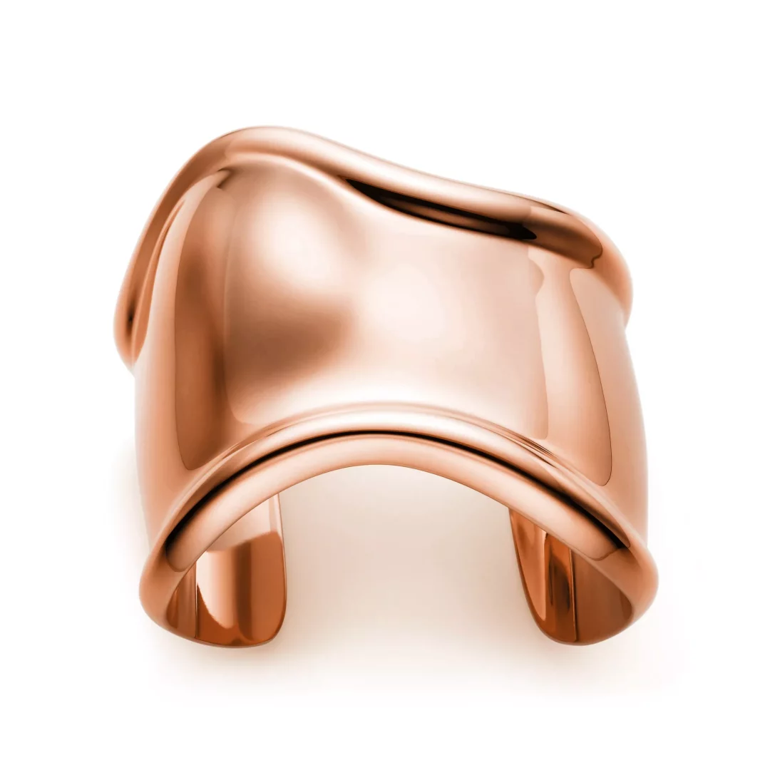 Beyaz bir arka plan üzerinde, Elsa Peretti'nin Tiffany için tasarladığı ikonik Bone Cuff koleksiyonuna ait, parlak, rose altın tonlarında, dalgalı ve heykelsi hatlara sahip, geniş, açık uçlu bir bilezik.