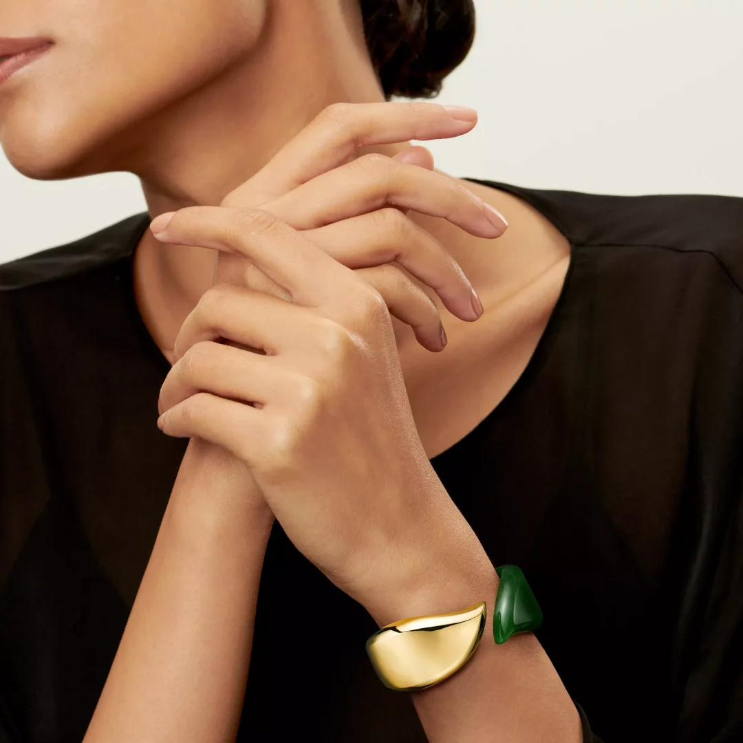 Bir kadının elleri zarifçe birleşmişken, sağ bileğinde Tiffany & Co.'nun ikonik Elsa Peretti Bone Cuff tasarımına sahip, parlak altın rengi ve zümrüt yeşili bölümlerden oluşan, heykelsi ve akıcı hatlara sahip, modern bir bileklik görülüyor.