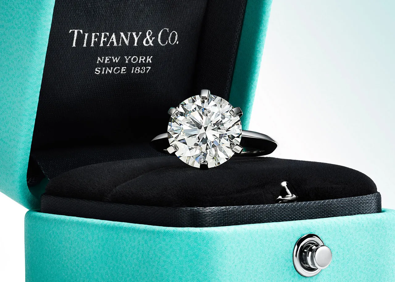 Açık duran ikonik Tiffany mavisi kutunun içinde, siyah kadife zeminde altı tırnaklı gümüş montürüyle büyük, parlak kesim bir pırlanta yüzük zarifçe duruyor ve kapağında Tiffany & Co. logosu belirgin bir şekilde görülüyor.