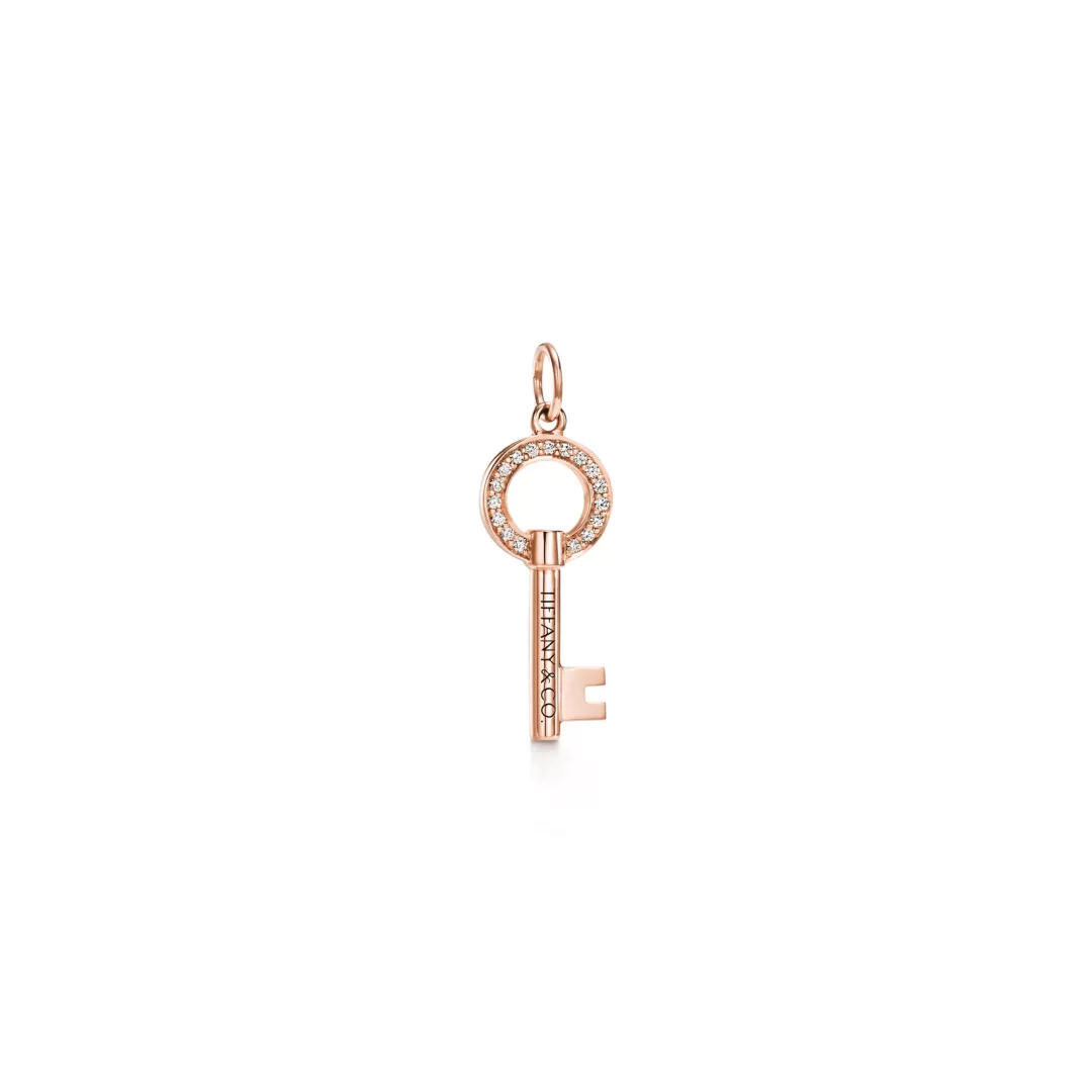 Tiffany & Co. markasının özel Tiffany Keys koleksiyonundan, pırlanta taşlarla çevrelenmiş yuvarlak anahtar başlığına ve sapında markanın adının yazılı olduğu, ışıltılı pembe altından üretilmiş zarif bir anahtar kolye ucu, sade beyaz bir arka planda sergileniyor.