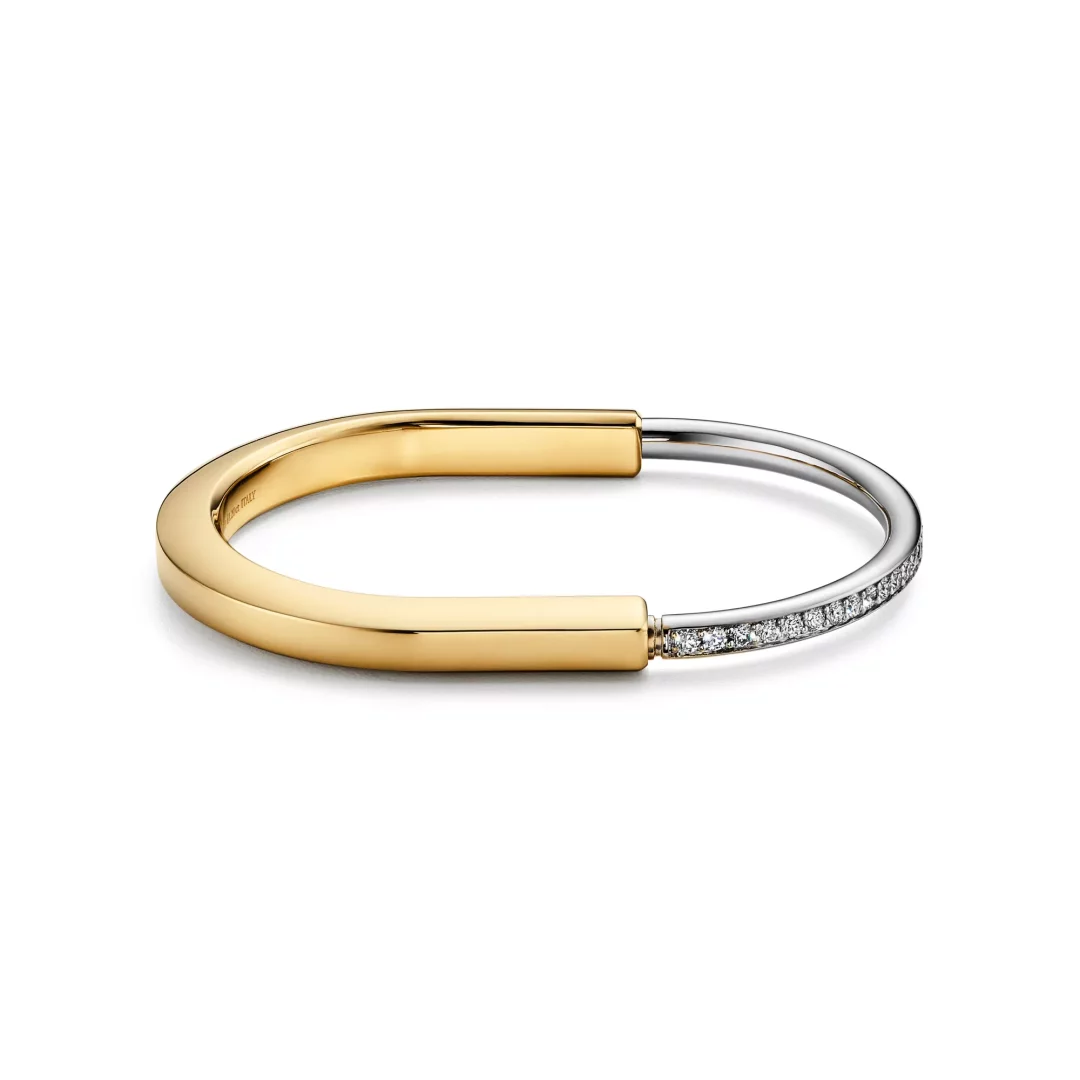 Tiffany Lock koleksiyonuna ait, parlın yüzeyli kalın, U şeklinde sarı altın bir bölümün, pırlantalarla bezenmiş zarif beyaz altın bir bantla birleştiği, modern tasarımlı, iki renkli bir bileklik beyaz arka plan üzerinde dikkat çekici bir şekilde sergileniyor.