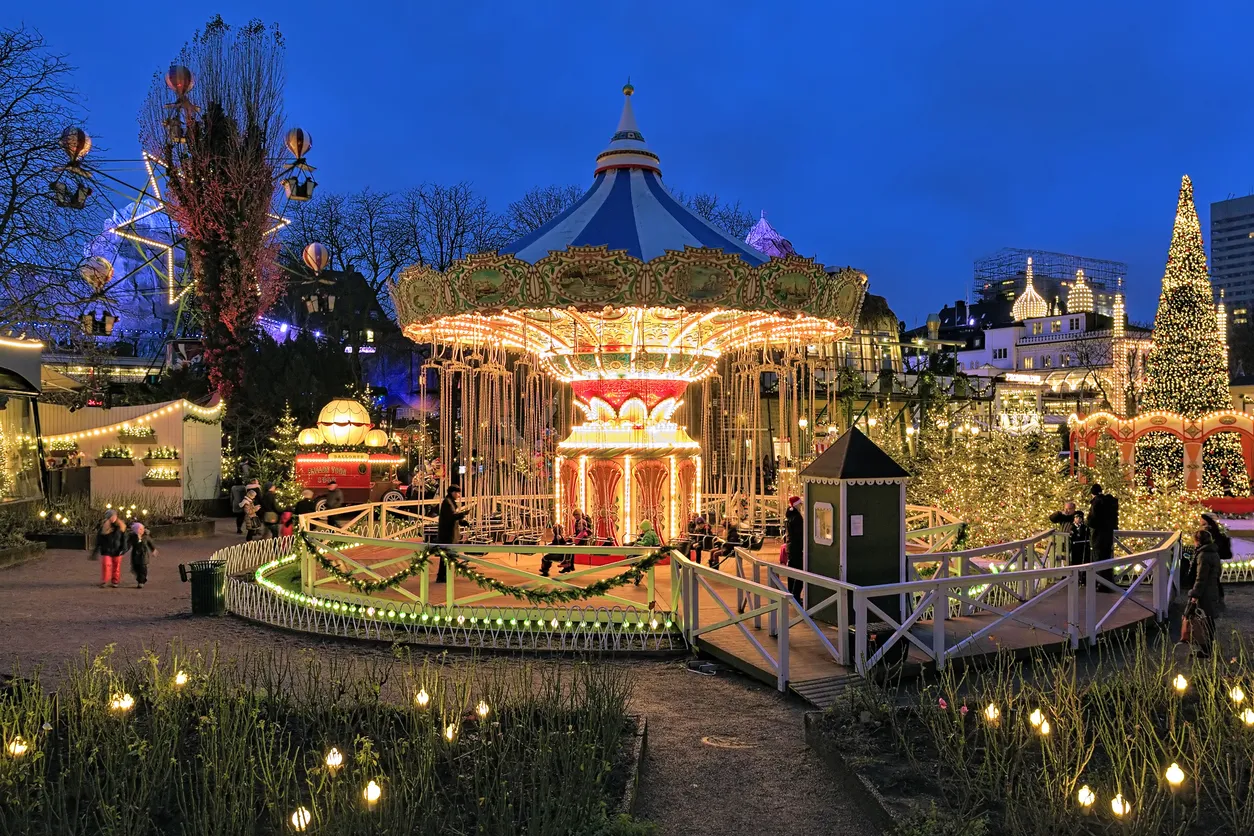 Alacakaranlıkta, Tivoli'deki canlı bir Noel pazarında, ışıl ışıl parlayan zincir dönme salıncak ve yıldız şeklinde dönme dolabın yanı sıra dev bir Noel ağacı, etraftaki diğer süslemeler ve ziyaretçilerle birlikte büyülü bir kış festivali atmosferi oluşturuyor.