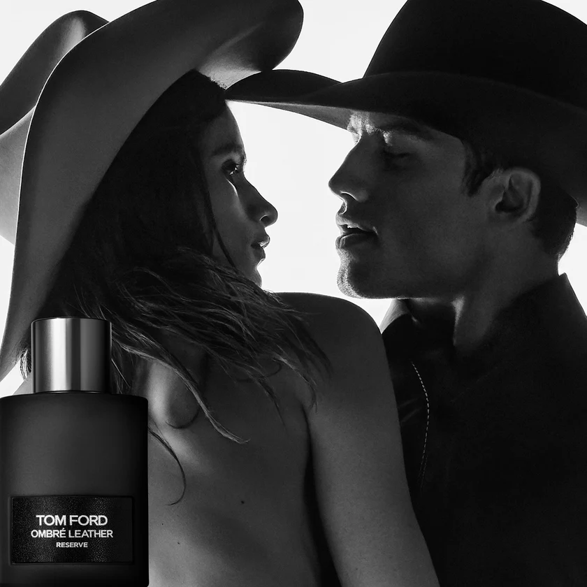 Tom Ford Ombré Leather Reserve parfüm şişesi, kovboy şapkası takan bir kadın ve erkeğin birbirine yakın, dramatik ışıklarla aydınlatılmış, samimi siyah beyaz portresinin ön planında dikkat çekiyor.