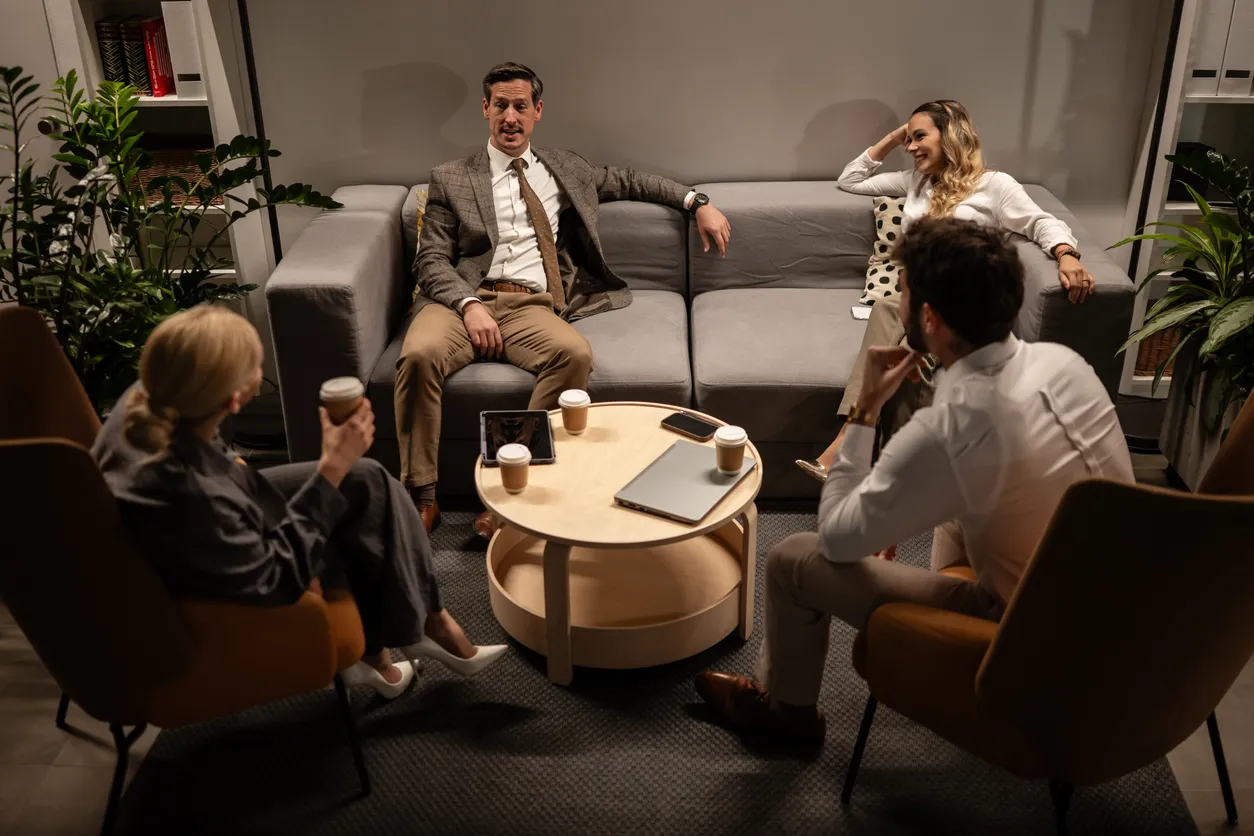 Modern bir ofis lounge alanında, gri bir kanepe ve iki kahverengi koltukta oturan dört iş arkadaşı, yuvarlak ahşap sehpada duran kahveler, tablet, akıllı telefon ve laptop eşliğinde samimi bir iş toplantısı gerçekleştiriyor, bu görüntü iş hayatındaki verimli ekip toplantılarının rahat bir ortamda da yapılabileceğini vurguluyor.