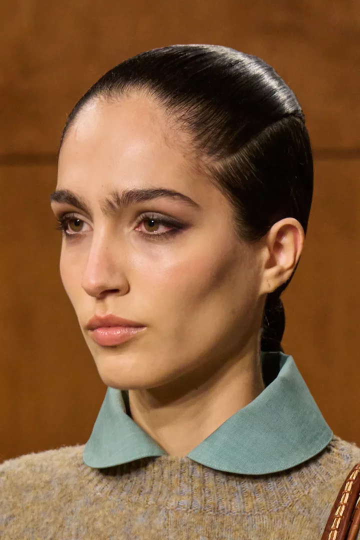 Modelin yakın çekim portresinde, Tory Burch Sonbahar 2026 Ready-to-Wear defilesinden, doğal kalın kaşları, belirgin eyeliner ve parlak nude dudak makyajı dikkat çekiyor; saçları parlak ve arkaya taranmış, kulak arkasında zarif bir örgüyle toplanmış, üzerinde sade örgü bir kazak ve mint yeşili yuvarlak yakalı bir gömlek görülüyor.
