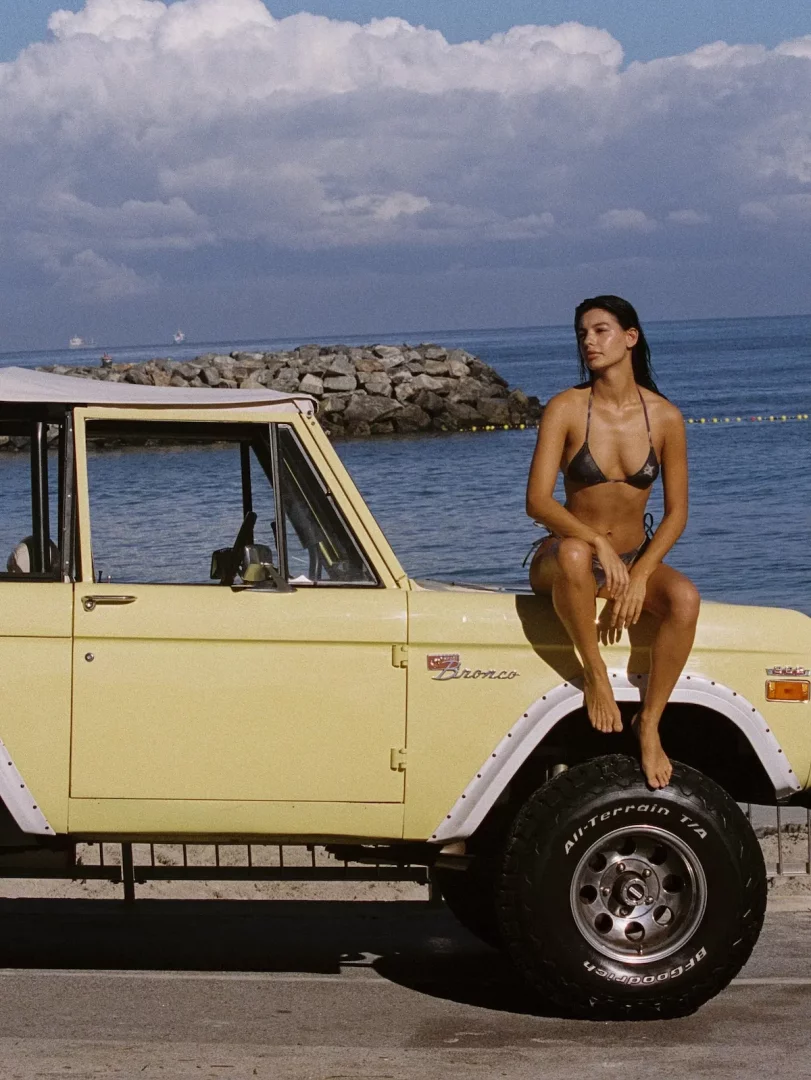 Parlak, desenli bir bikini mayo giyen, üstü açık sarı bir Ford Bronco'nun kaputu üzerine oturmuş genç bir kadın, arkasında masmavi deniz, kayalıklar ve bulutlu gökyüzüyle güneşli bir sahilde.