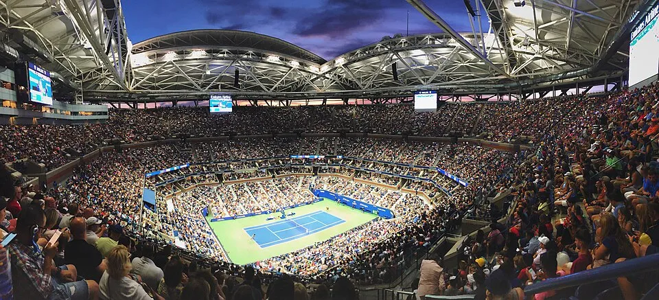 Amerika Açık tenis turnuvası sırasında, çatısı açık büyük bir stadyumda, gece mavisine dönen gökyüzünün altında, korttaki iki tenisçiyi izleyen binlerce seyirci tribünleri dolduruyor ve parlak ışıklar sahayı aydınlatıyor.