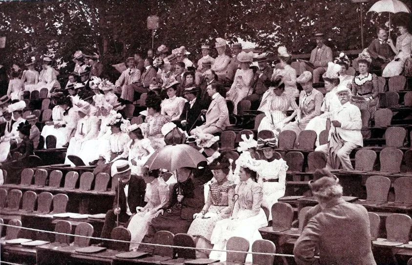 1900'lü yılların başlarında gerçekleşen US Open tenis turnuvasının tarihçesi anlarından birini yansıtan bu geniş açılı görselde, şık elbiseler, büyük şapkalar ve takım elbiselerle dönemin modasını sergileyen yüzlerce seyirci, kalabalık tribünlerde otururken güneşten korunmak için bazıları şemsiye kullanıyor.