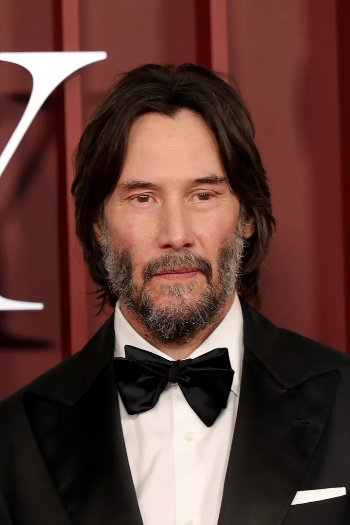 Keanu Reeves, omuzlarına dökülen koyu renk dalgalı uzun saçları ve gür, grileşmiş sakalıyla, siyah smokini ve papyonu içinde kırmızımsı fon önünde objektife bakarken modern bir uzun erkek saç modeli sunuyor.