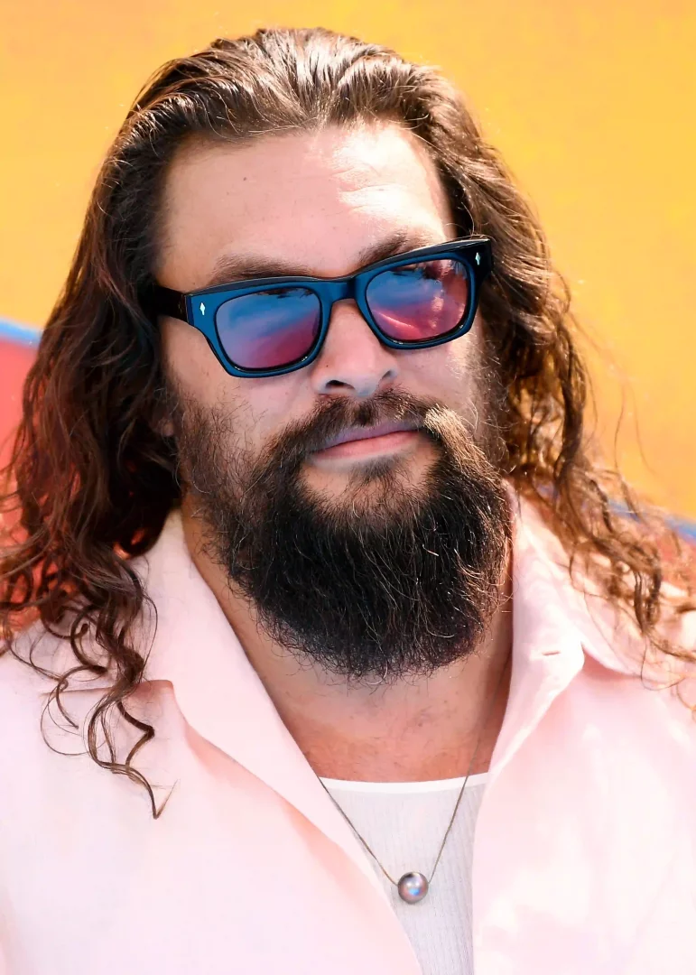 Güneş gözlüklü, gür sakallı ve dalgalı uzun saçları omuzlarına dökülen Jason Momoa, pembe gömleği ve tek inci kolyeli beyaz tişörtüyle, uzun erkek saç modellerine uygun modern bir görünüm sergiliyor.