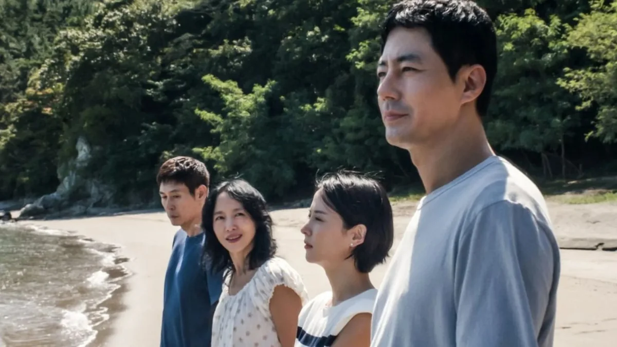 Possible Love filminden bir karede, denizin dalgalı kıyısında duran dört karakter, en sağdaki adamın ileriye dönük dalgın bakışı ve diğerlerinin farklı yönlere yönelmiş yüz ifadeleriyle, arka plandaki yeşil ağaçlarla çevrili sakin anı yansıtıyor.