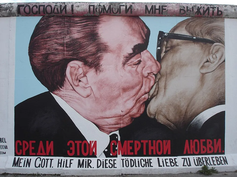 Berlin Duvarı üzerinde yer alan ikonik bir grafiti resminde, soluk mavi bir fonun önünde, koyu renk takım elbiseli ve beyaz gömlekli iki adamın dudak dudağa öpüşen yakın çekim portresi belirgin şekilde gösteriliyor; Rusça ve Almanca dillerinde Tanrım, bu ölümcül aşkta hayatta kalmama yardım et anlamı taşıyan mesajlar, eserin siyasi ve tarihi önemini vurguluyor.
