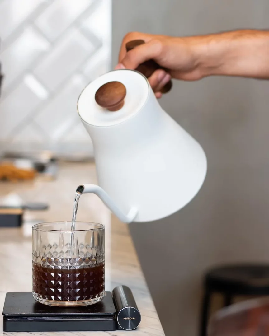 Beyaz, ahşap detaylı gooseneck kettle'dan, şeffaf, desenli cam bir bardağın içindeki koyu renkli kahveye hassas bir şekilde su akıtılıyor; bu özenli kahve hazırlığı Hirdia markalı dijital bir tartının üzerinde gerçekleşiyor.