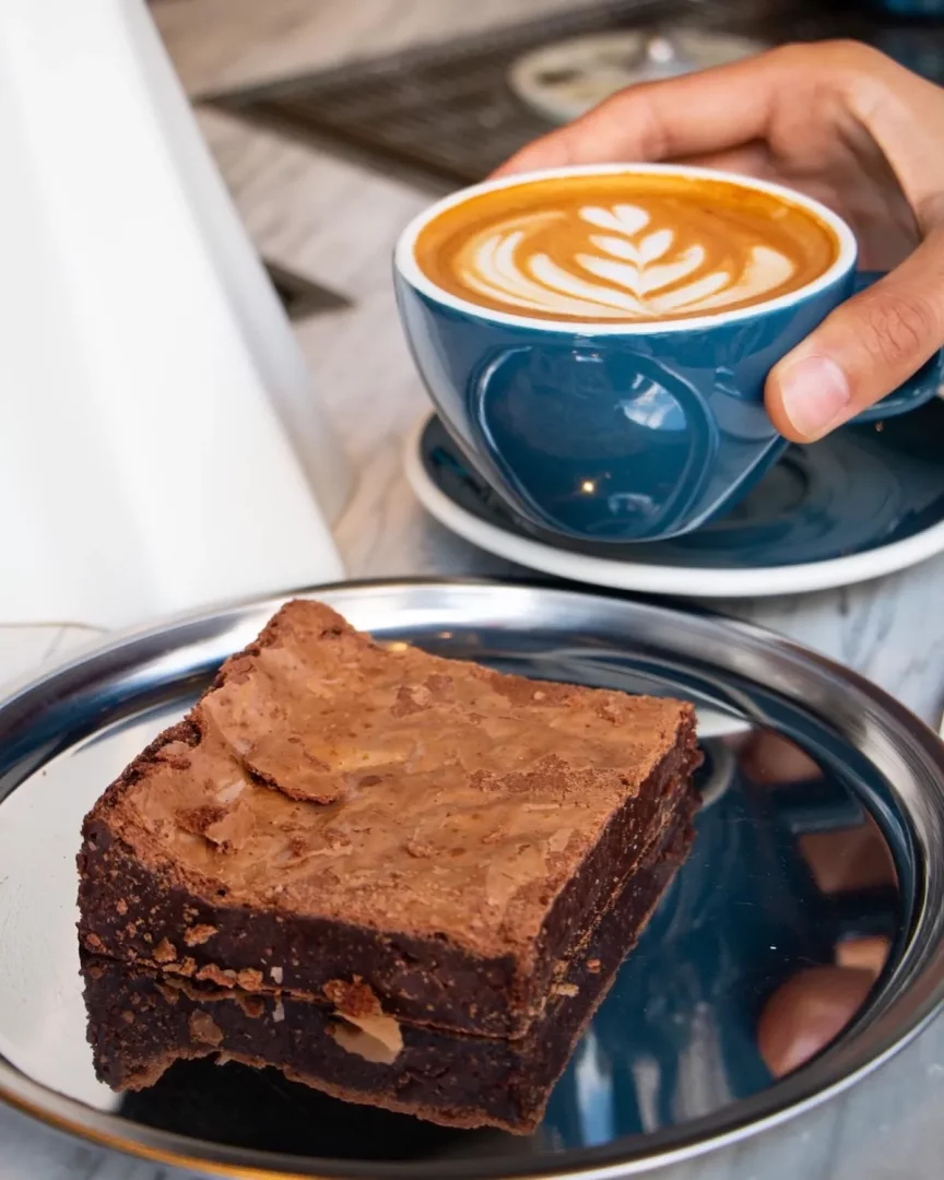 Mavi bir fincanda, üzerinde zarif bir latte sanatı bulunan taze demlenmiş kahveyi tutan bir el, yanında parlak gümüş bir tepside servis edilen, ceviz parçacıklı ve yoğun çikolatalı bir brownie ile Williams Roastery atmosferindeki keyifli bir anı yansıtıyor.