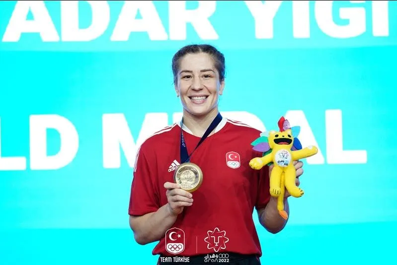 Yasemin Adar Yiğit, milli takım kırmızı formasını giyerek boynundaki altın madalyayı sağ eliyle tutarken sol elinde Oran 2022 Akdeniz Oyunları maskotunu gururla sergiliyor, mavi bir zemin ve beyaz harfler önünde zaferini kutluyor.