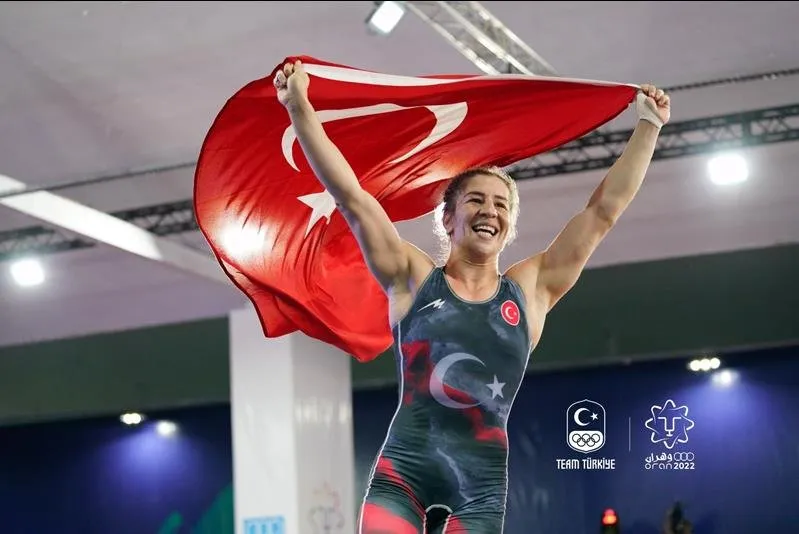 Yasemin Adar Yiğit, Oran 2022'deki başarısının ardından kollarını havaya kaldırarak dalgalandırdığı büyük Türk bayrağının altında gururla gülümsüyor, koyu renkli güreş mayosunda da ay yıldız deseni belirginleşiyor.
