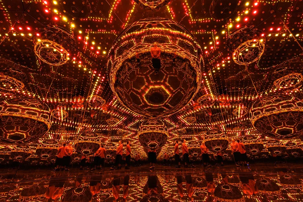 Yayoi Kusama'nın bir Sonsuzluk Odası eserinde, tavan ve duvarlardan yansıyan parlak turuncu, kırmızı ve sarı ışık noktacıklarıyla dolu aynalı bir odada, zeminde yansımalarıyla birlikte duran birkaç insan figürü ve ortada asılı duran, çok sayıda altıgen yüzeye sahip büyük, yansıtıcı kürelerin sonsuz yansımaları görülüyor.