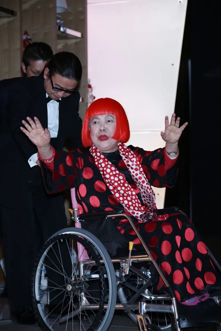Kırmızı puantiyeli siyah elbisesi ve canlı kızıl saçlarıyla tanınan Yayoi Kusama, pembe tekerlekli sandalyesinde otururken ellerini havada coşkuyla sallıyor; arkasında smokinli bir adamın durduğu bu kare, sanatçının hastalığına rağmen enerjisini ve ikonik tarzını gözler önüne seriyor.