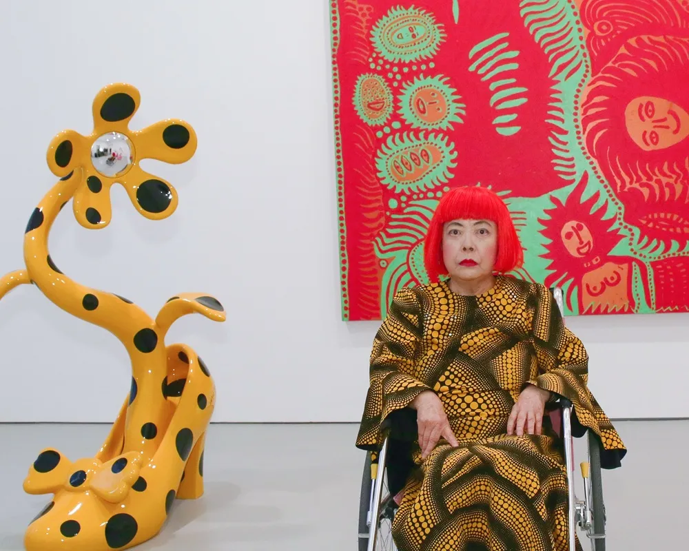 Parlak kırmızı saçlı ve puantiyeli desenli elbisesiyle tekerlekli sandalyede oturan sanatçı Yayoi Kusama, arkasındaki kırmızı-yeşil soyut tablo ve yanındaki sarı puantiyeli ayakkabı-çiçek heykeli gibi kendine özgü eserleri arasında poz veriyor.