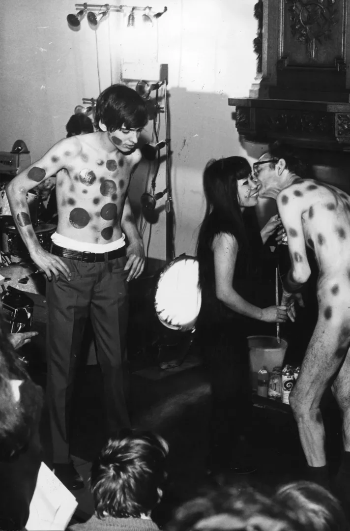 Yayoi Kusama'nın kim olduğunu anlatan bu siyah beyaz fotoğrafta, sanatçının imzası beneklerle boyanmış, pantolonlu üstsüz bir adamın solda durduğu, ortadaki uzun saçlı kadının yanağını öpen yarı çıplak başka bir adamın sağda yer aldığı, seyircilerin de izlediği bir performans sanatı sahnesi yansıyor.