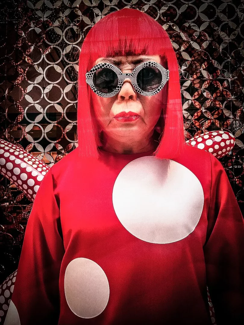 Kırmızı bob kesim peruklu, siyah puantiyeli dev gözlüklü ve iki büyük beyaz puantiyeli kırmızı bluz giymiş Yayoi Kusama'nın karakteristik bir figürü, arkasında dairesel motiflerle bezeli parlak bir arka plan ve puantiyeli heykellerle birlikte duruyor.