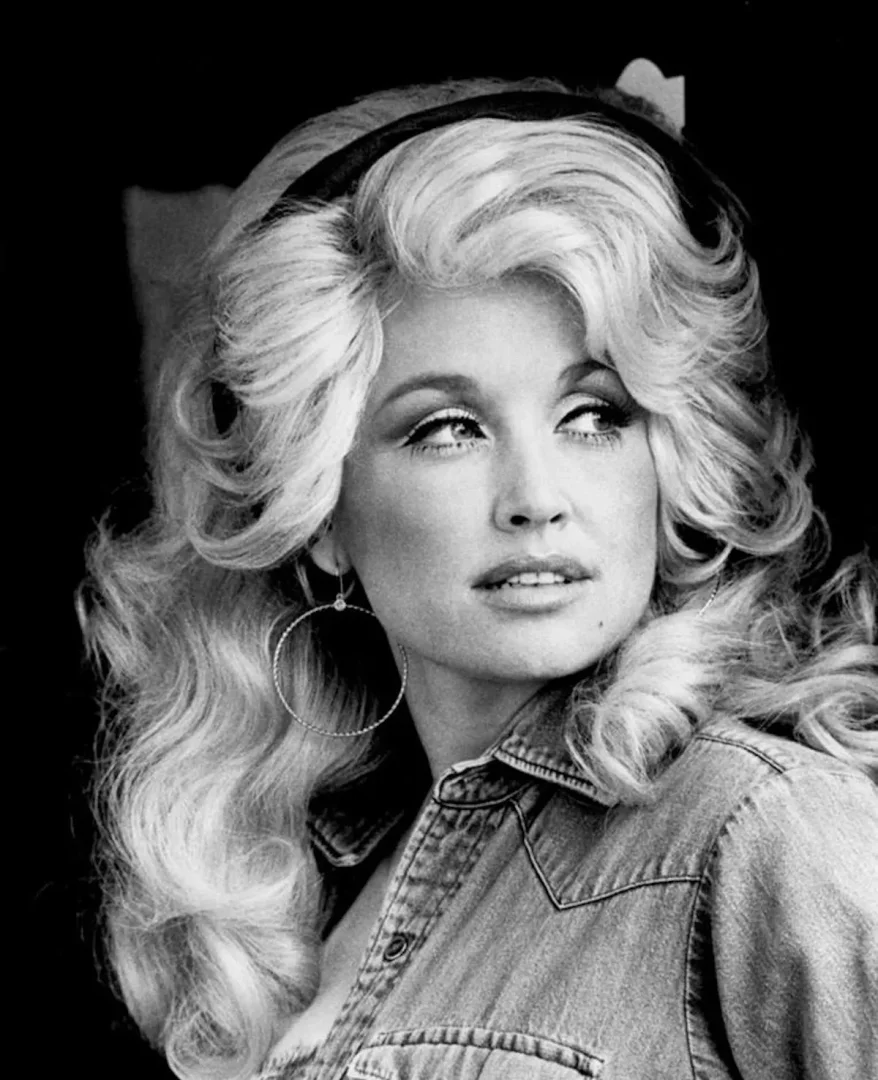 Bu siyah beyaz portrede genç Dolly Parton, meşhur hacimli sarı saçları, koyu bir saç bandı, büyük halka küpeleri ve denim gömleğiyle hafifçe yana dönmüş, uzaklara bakarken düşünceli ve zarif bir ifade sergiliyor.