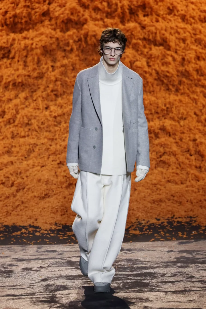 Podyumda yürüyen bir model, açık gri yün blazer ceketini, krem rengi balıkçı yaka kazağını, aynı renk bol kesim pantolonunu ve eldivenlerini gözlükleriyle tamamlamış, turuncu dokulu bir arka plan önünde şık bir kış stilini sergiliyor.