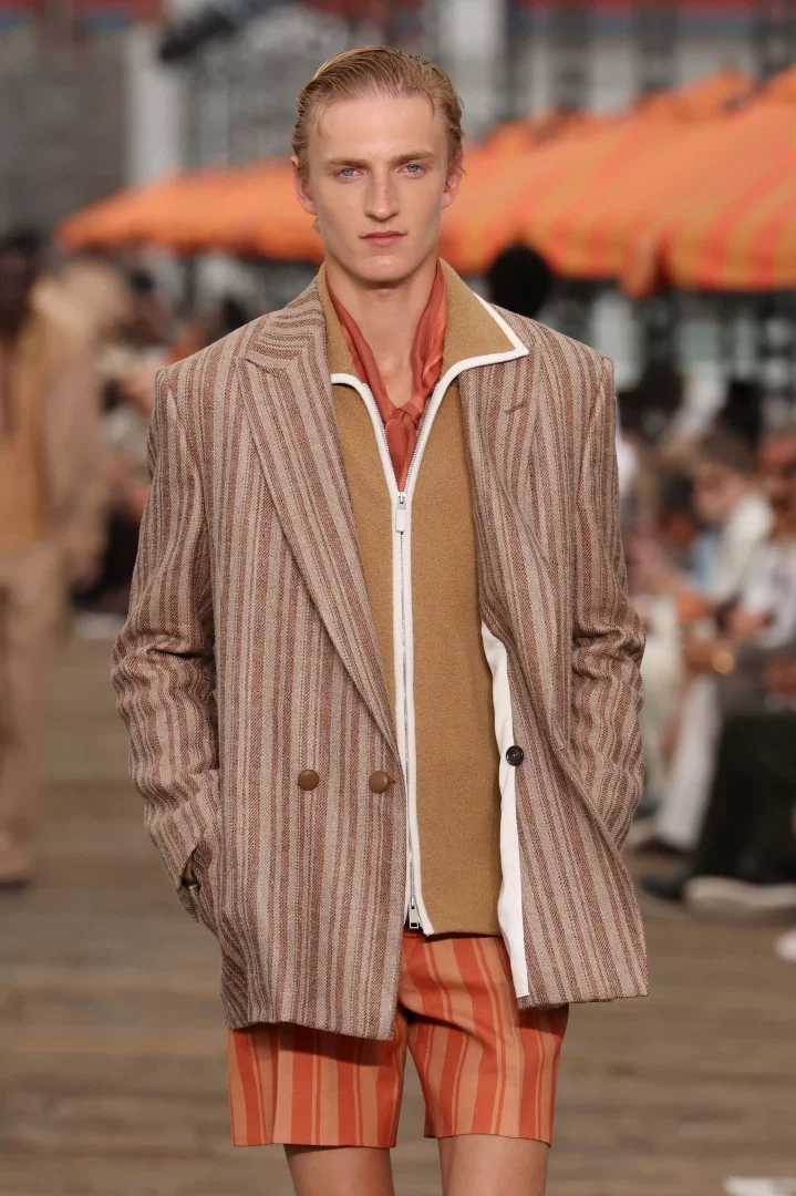 Açık renk saçlı, genç bir erkek model, kahverengi tonlarında çizgili çift düğmeli bir Zegna blazer ceket, içine giydiği bej renk fermuarlı bir üst ve turuncu çizgili şortuyla podyumda ciddi bir ifadeyle yürüyor.