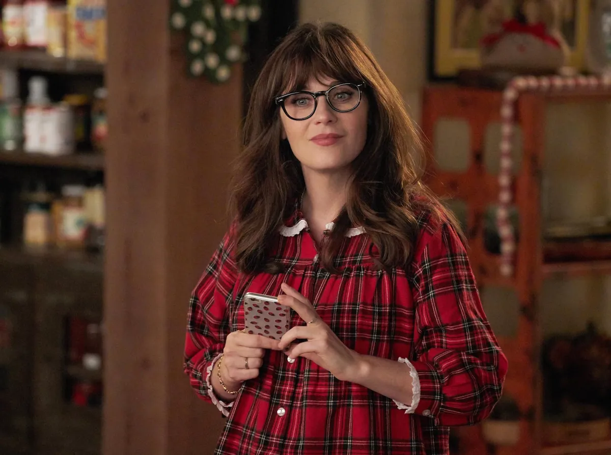 Zooey Deschanel, New Girl dizisindeki Jess karakteri olarak, yanakları hafifçe kızarmış, yuvarlak çerçeveli gözlükleri ve beyaz yakalı kırmızı ekoseli pijamasıyla rahat bir ev ortamında, desenli cep telefonunu iki eliyle tutarken gülümseyerek kameraya bakıyor.