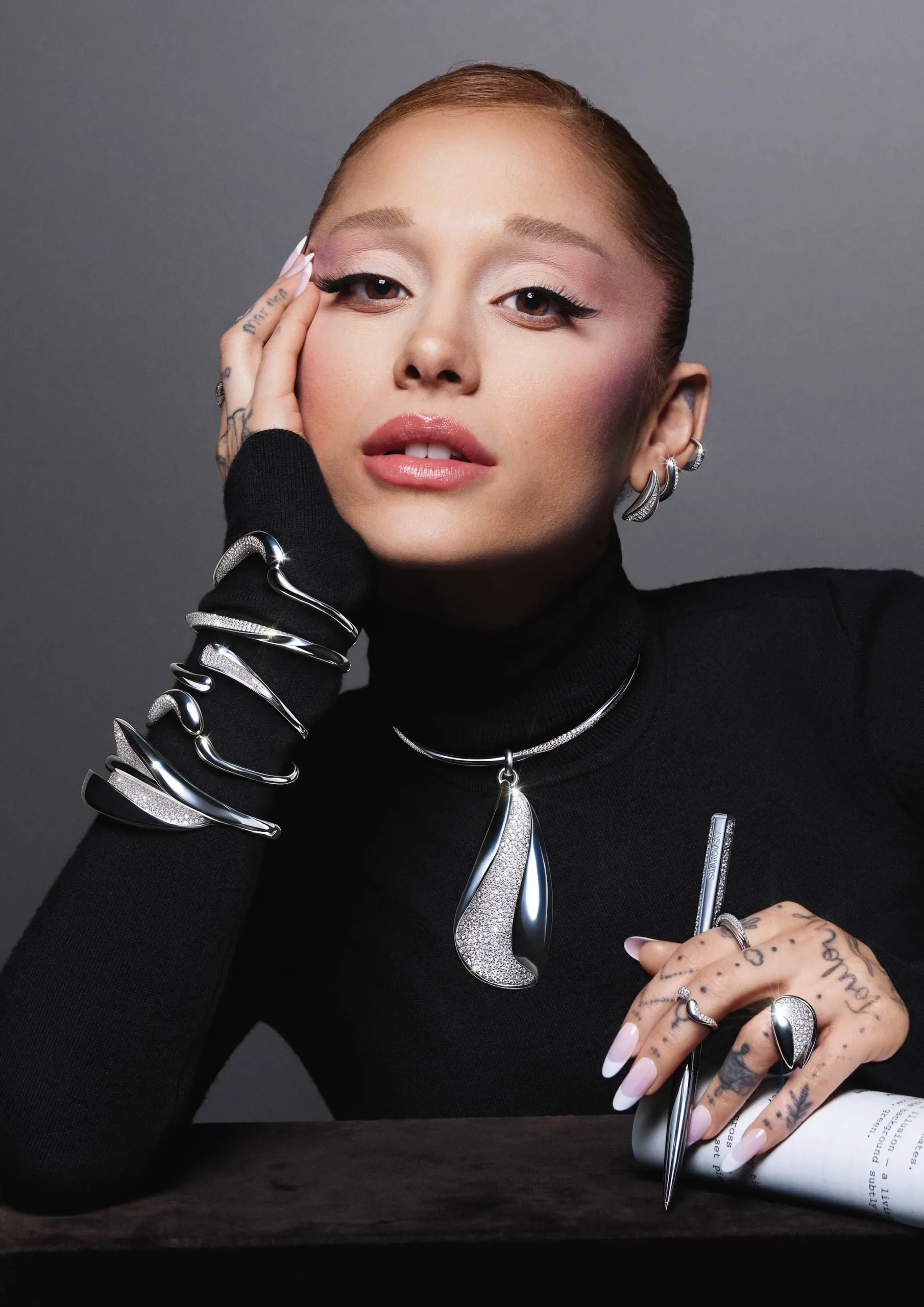 Siyah boğazlı bir kazak giyen Ariana Grande, zarifçe elini yüzüne dayamış, parıltılı kristal taşlarla süslü gümüş renkli Classica takılarını ve aynı tasarıma sahip bir Swarovski kalemini kameraya bakarak sergiliyor.