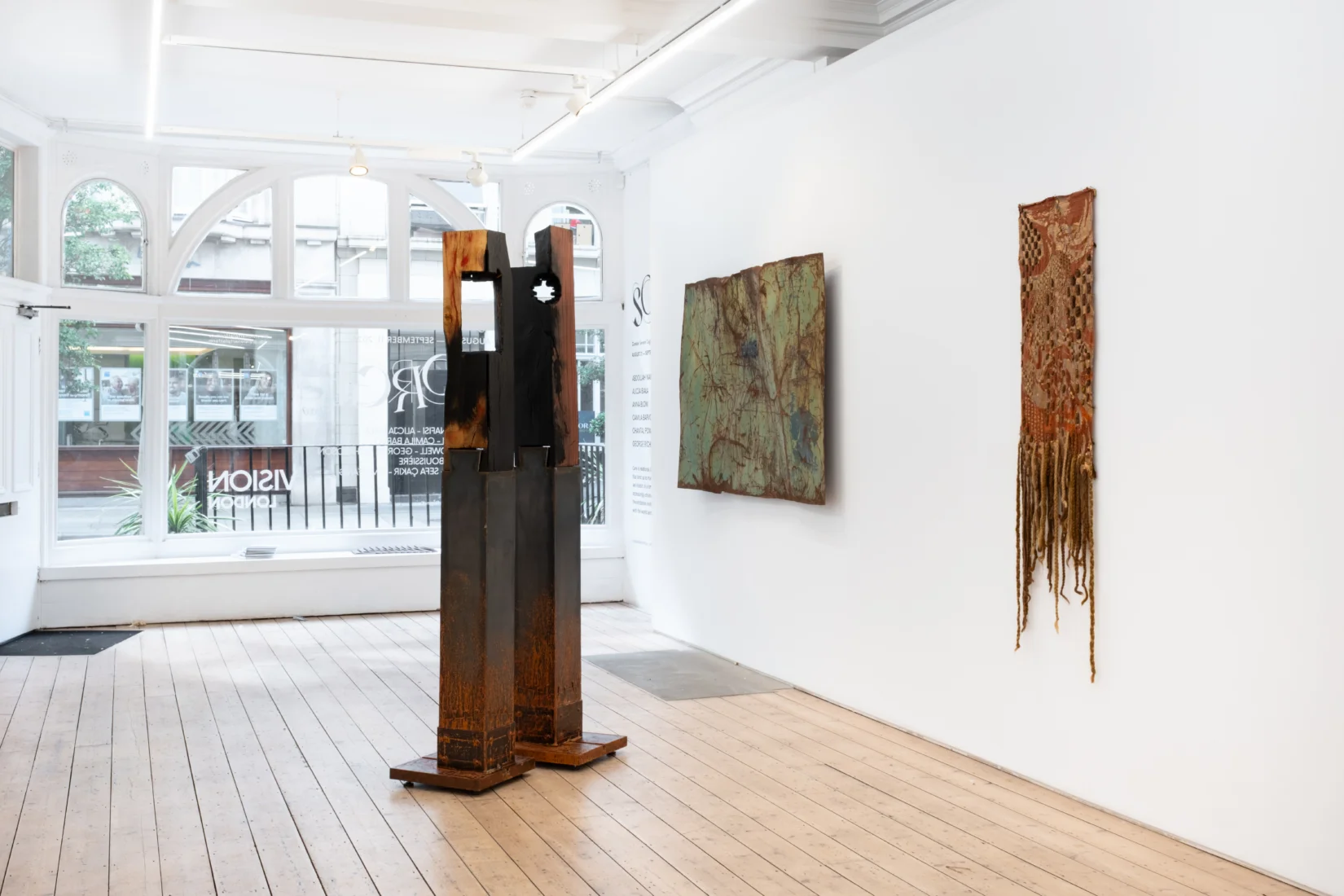 Beyaz duvarlı modern bir sanat galerisinde, Vision Art Platform'un Lightscapes sergisinden Charles-Joseph de Ligne'in büyük, yansıtıcı ve koyu renkli fotoğrafına sırtı dönük bakan bir izleyici, eserin parlak yüzeyinde yansıyan çevresiyle birleşerek farklı bir bakış deneyimliyor.