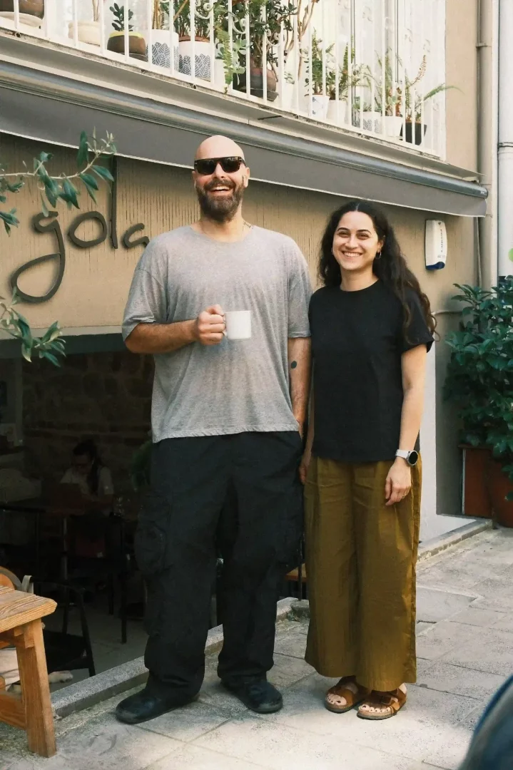 Gola restoranının önünde, incir mevsimi lezzetlerini menülerine taşıyan şefler Yılmaz Tüleyli ve Ezgi Yıldırım, Yılmaz'ın siyah güneş gözlükleri ve elinde tuttuğu beyaz kupasıyla, Ezgi'nin ise uzun dalgalı saçları ve içten gülümsemesiyle yan yana poz veriyor.