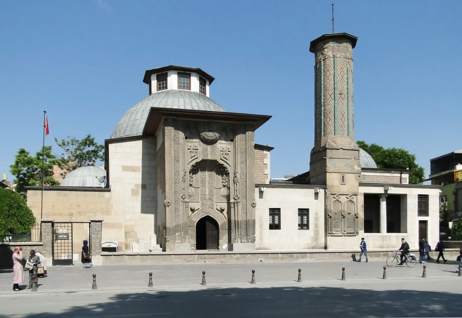 Parlak mavi gökyüzü altında, Konya'daki Selçuklu döneminden kalma İnce Minareli Medrese, detaylı taş oymalı taç kapısı ve geometrik desenli çinilerle süslenmiş uzun minaresi ile cadde üzerindeki yayalar ve bisikletli bir kişiyle birlikte güneşli bir günde tüm ihtişamıyla görülüyor.