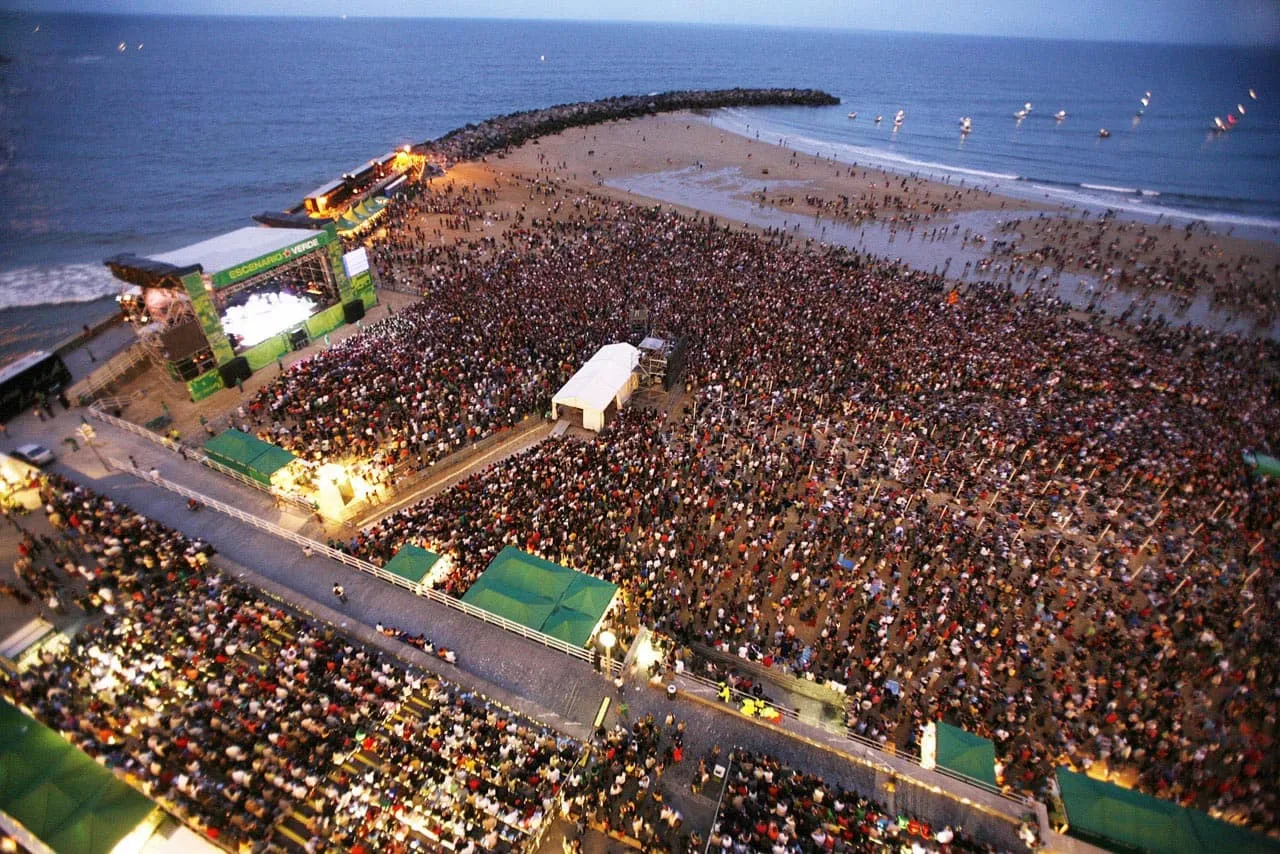 Bir Jazzaldia konserinde canlı bir atmosferde sahnedeki müzisyenler davul ve diğer enstrümanlarıyla coşkulu bir performans sergilerken, seyirciler sahne ışıklarının aydınlattığı geniş salonda müziğin tadını çıkarıyor ve bu unutulmaz anlara tanıklık ediyor.